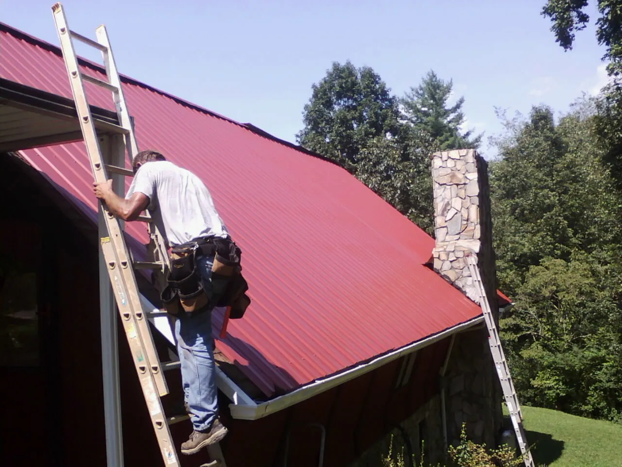 Roofer on a ladder beside a red metal roof and stone chimney — Soffit & Fascia Repair by Roofing Brothers in Joffre